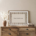 Framed artwork with 'Pour Forth Love' text on a wooden dresser against a beige wall.